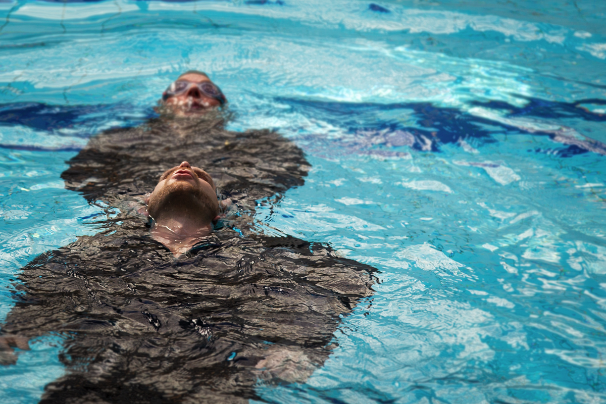 Två personer simmar på rygg där den ena personen övar på att rädda den andre ur vattnet i en simbassäng. 